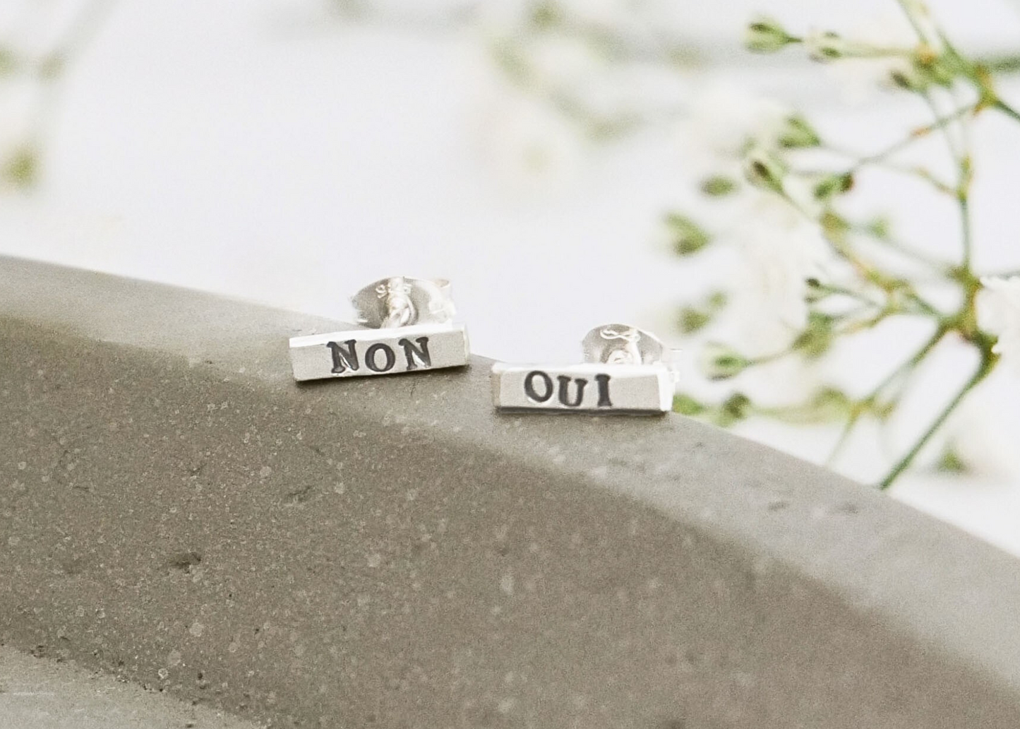 Silver stud earrings with 'NON' and 'OUI' on a stone surface with blurred greenery in the background